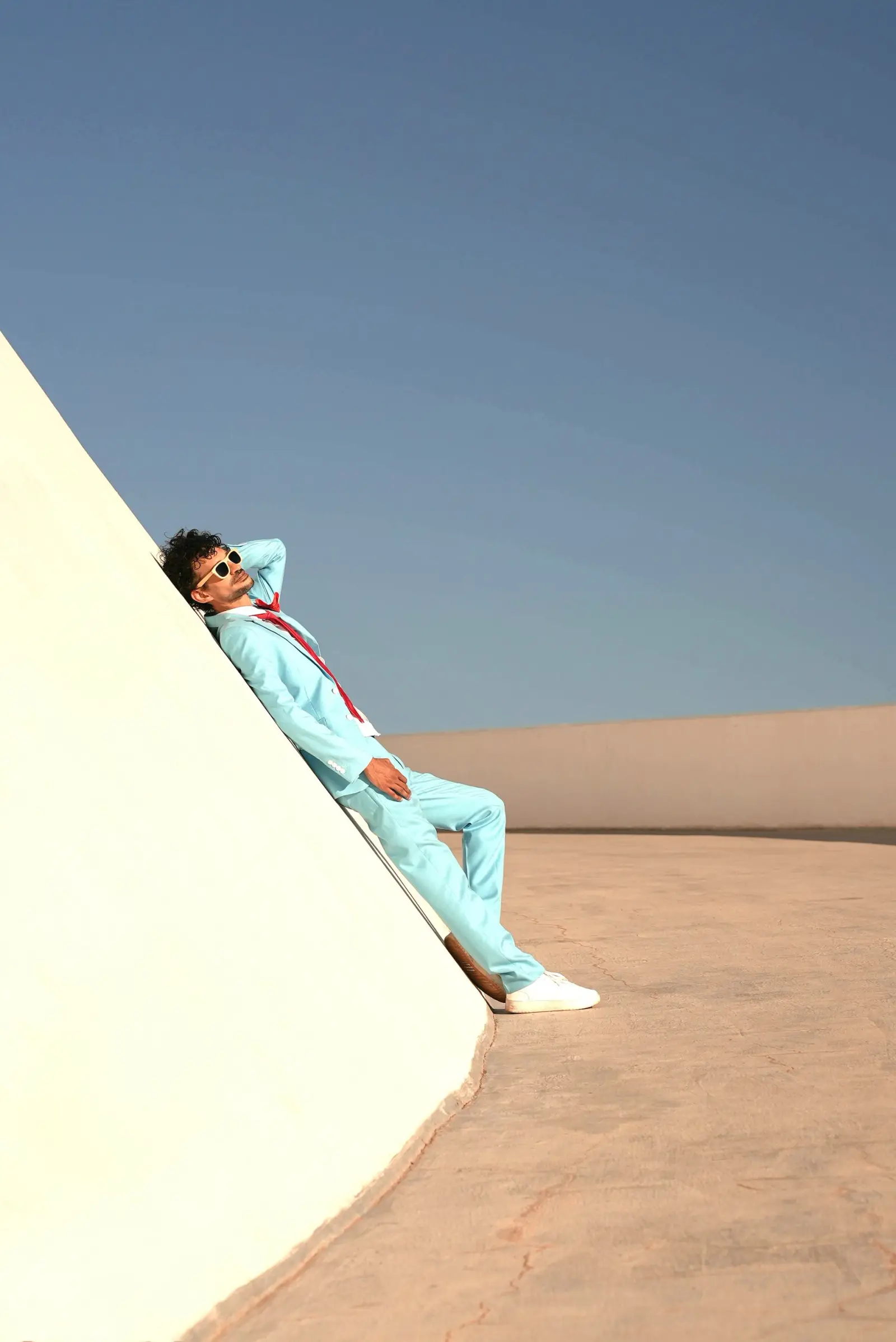 Marlon in a pale blue suit reclining against a curved white wall under a clear sky
